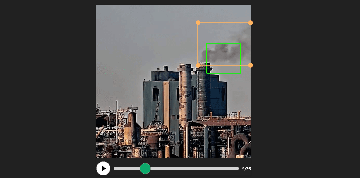 Foto van de Tata Steel fabriek. Er komt zwarte rook uit de schoorstenen. Om de rook is een oranje kader en een kleiner groen kader.