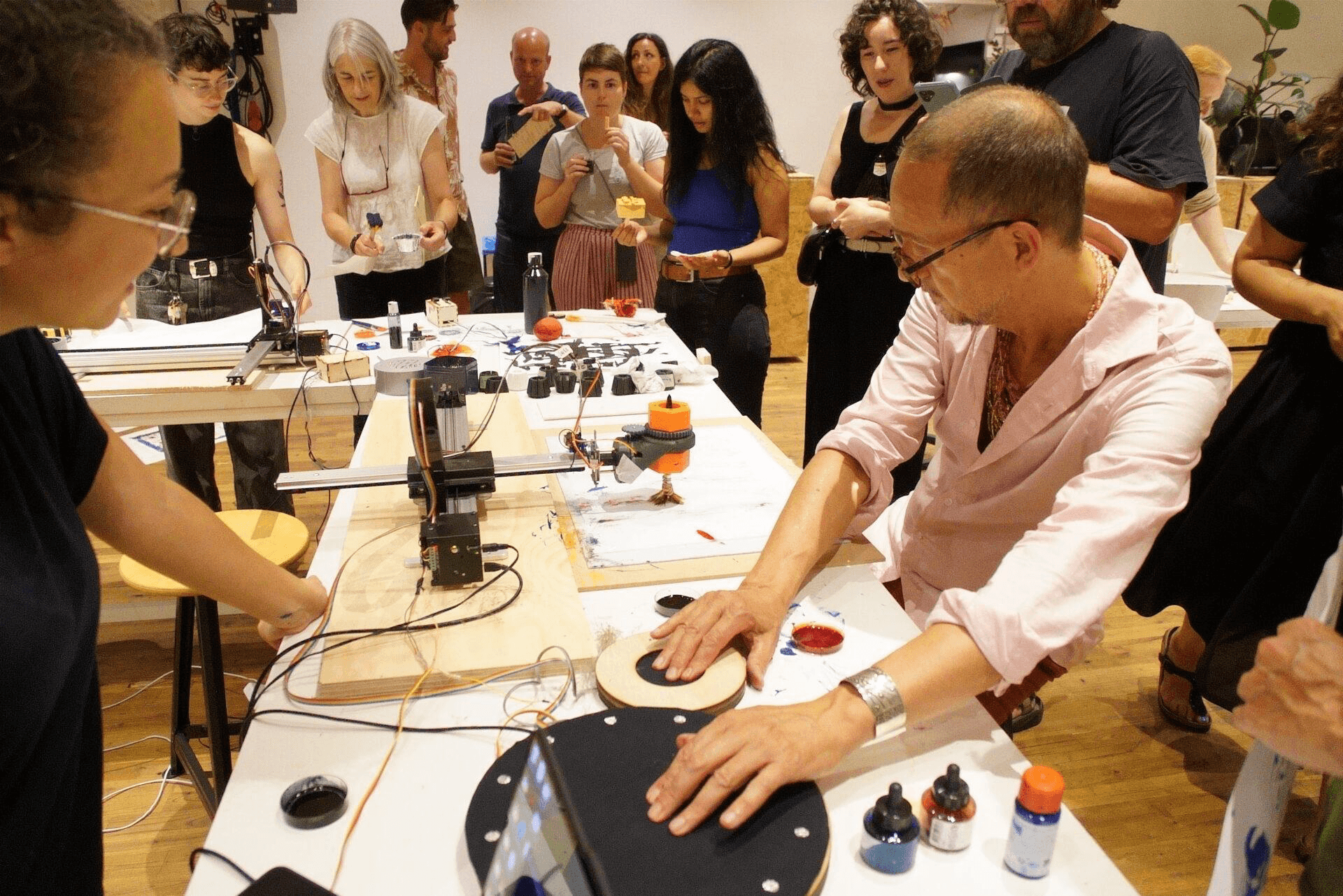 People are standing around a white table with several electronic tools on them. A man is touching a large black circlular touch pad and looking at a machine printing something.