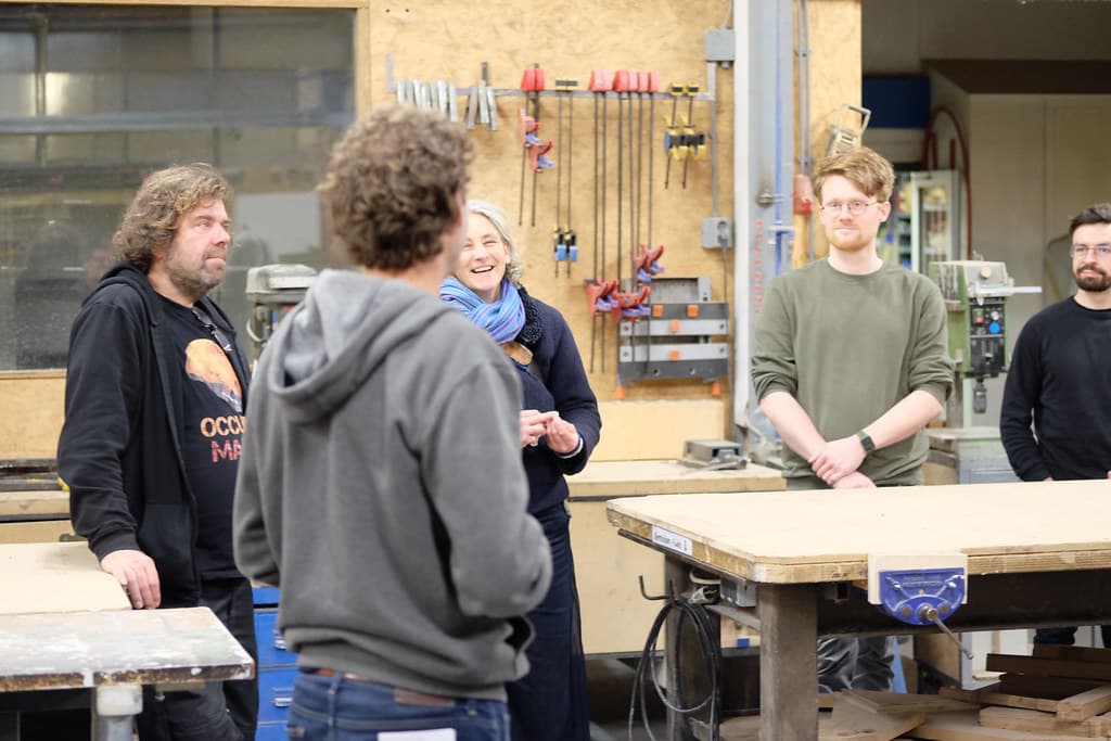 Foto van een groep mensen in een werkplaats. Een aantal mensen staan om een houten werktafel heen. Op een houten wand op de achtergrond hangen verschillende zagen.