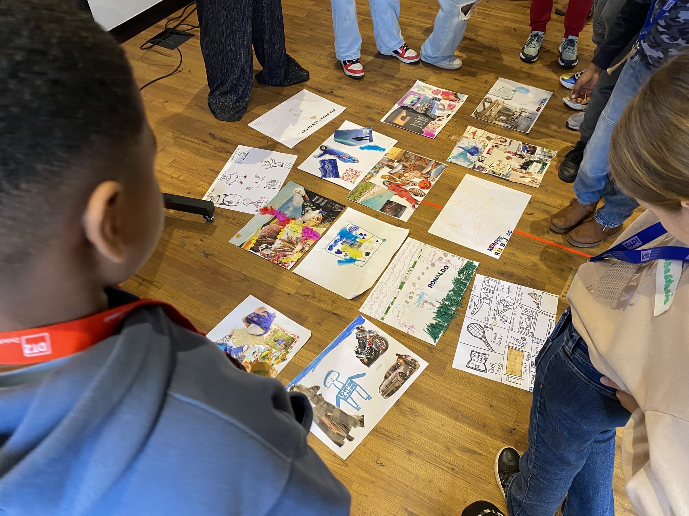 Kinderen staan om een houten tafelblad heen en bekijken A4'tjes met daarop hun tekeningen en concepten voor een kostumm gebaseerd op identiteit.