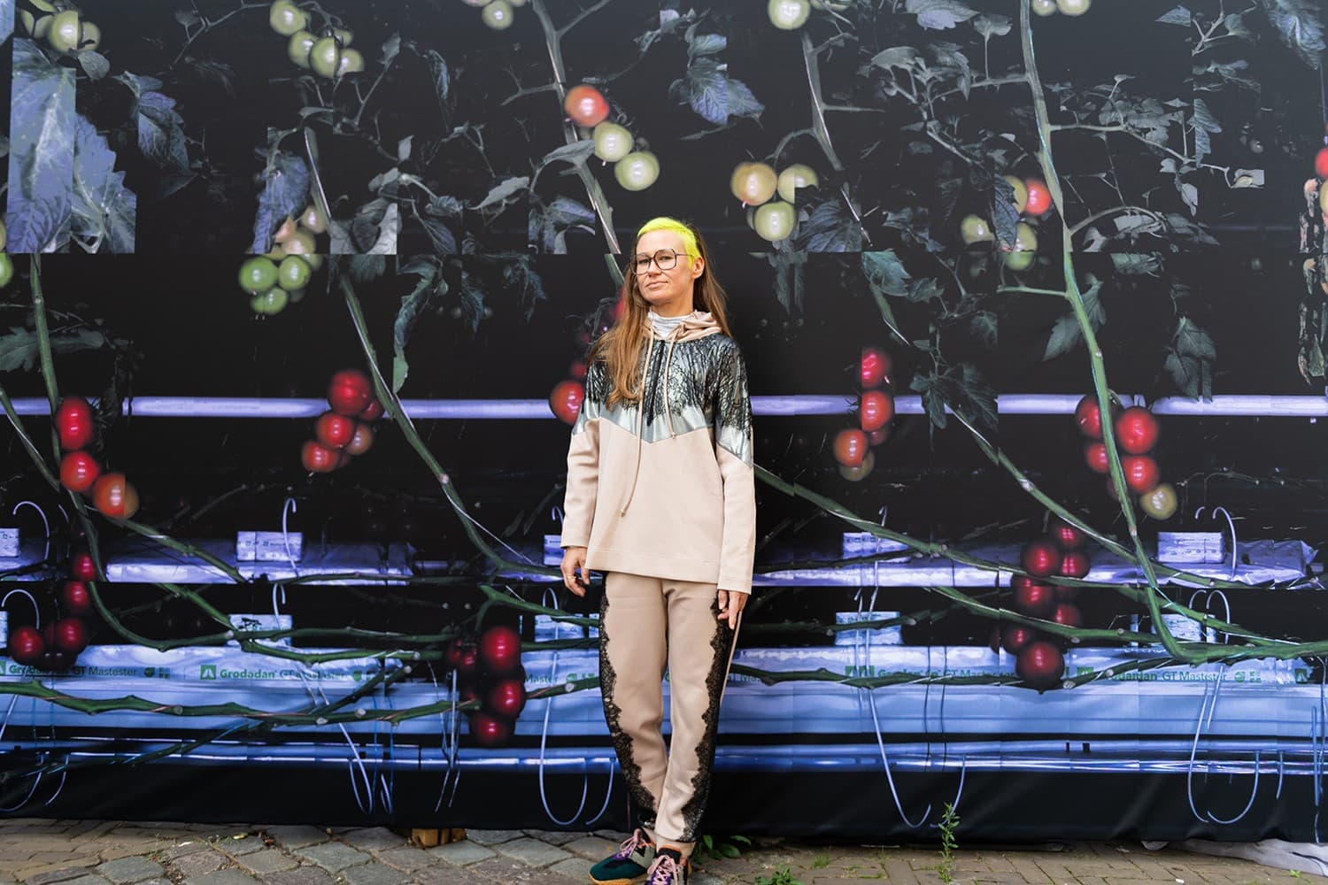 Artist Špela Petrič standing in front of her work, a big canvas of photos taken by a computer of tomato plants. Spela has long dark blonde hair, the side of her head has a buzz cut with neon yellow died hair.