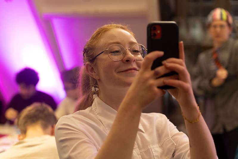 Een vrouw met een paardestaart en bril houd haar telefoon voor haar gezicht, haar telefoon omhoog gericht, alsof ze een foto neemt.
